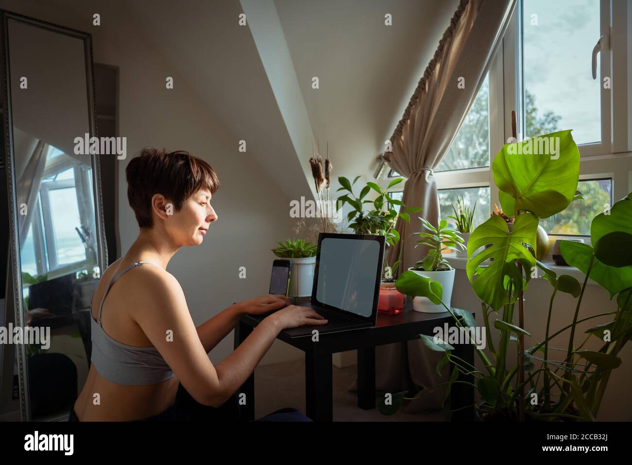 Femme de course mixte dans des vêtements de maison décontractés travaillant sur un ordinateur portable assis à une table basse avec beaucoup de plantes vertes en pot près de la fenêtre. Travailler à la maison Banque D'Images