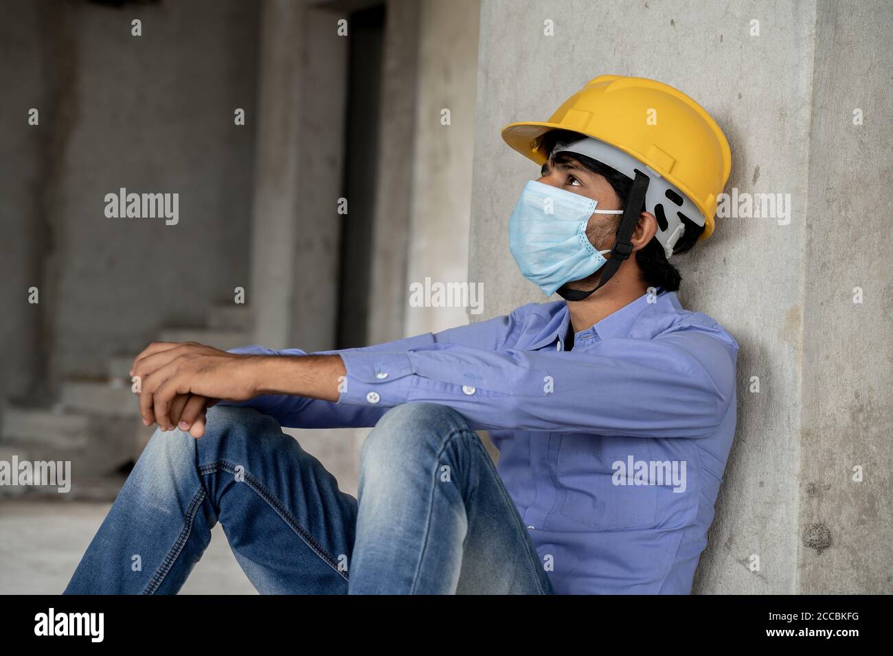 Vue latérale de la tête du travailleur de la construction qui voit à l'extérieur Bâtiment de construction fenêtre dans Sad - travailleur industriel dans un casque avec masque médical Banque D'Images
