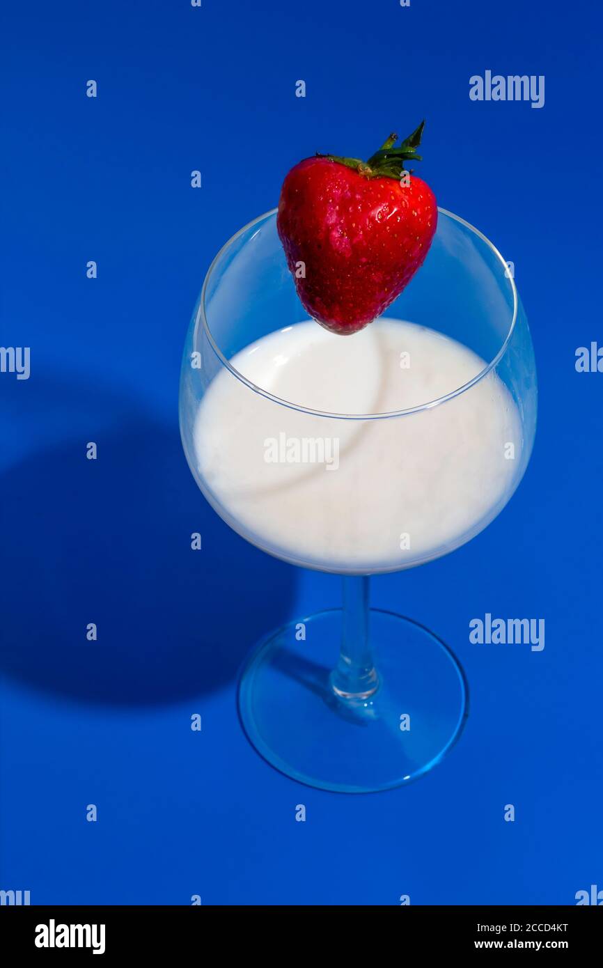 Fruits de fraise sur le point de tomber dans un verre de lait sur fond bleu Banque D'Images