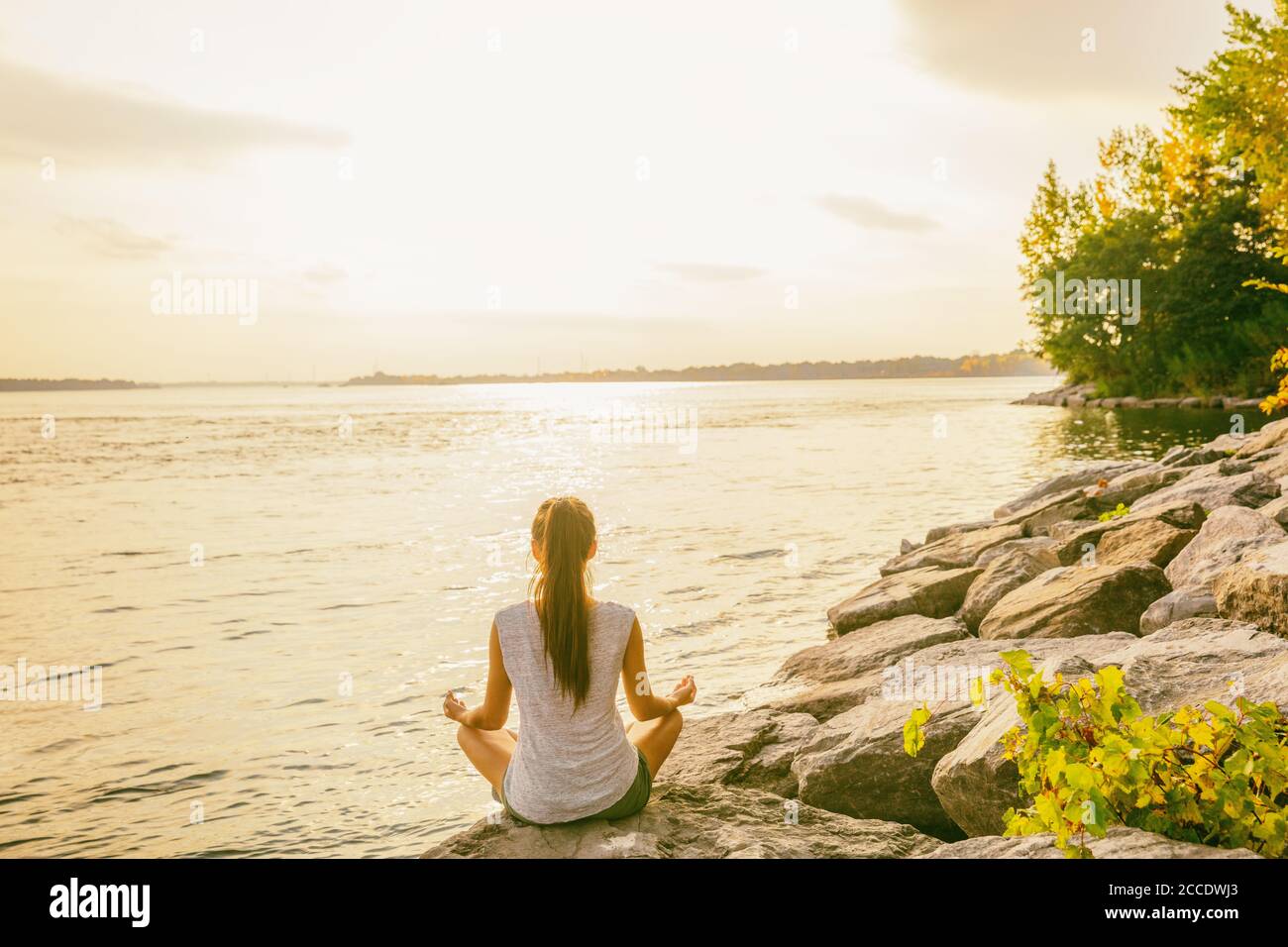 Cours de yoga à l'extérieur dans un parc naturel près du bord du lac. Une femme assise dans le lotus pose méditant par l'eau dans le soleil du matin se fait jour au lever du soleil. Méditation Banque D'Images