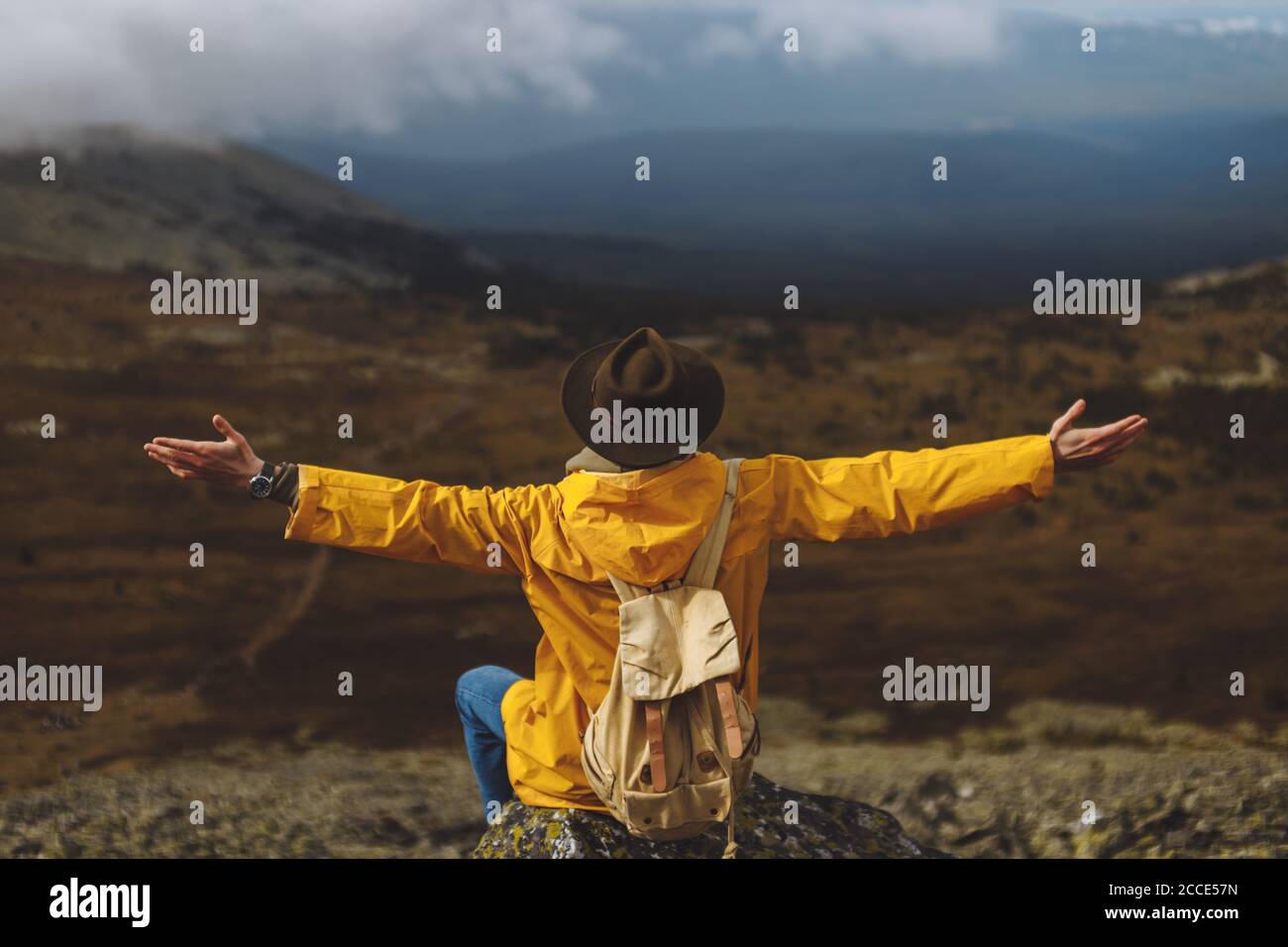 jeune touriste en manteau jaune et chapeau assis avec les bras tendus, saluant le nouveau jour, appréciant la vie. vue arrière photo Banque D'Images