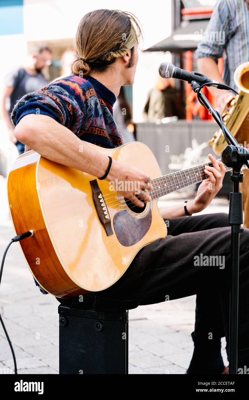 Photo verticale un jeune homme avec des cheveux longs attachés chantant et jouer une guitare dans la rue avec un saxophoniste Banque D'Images