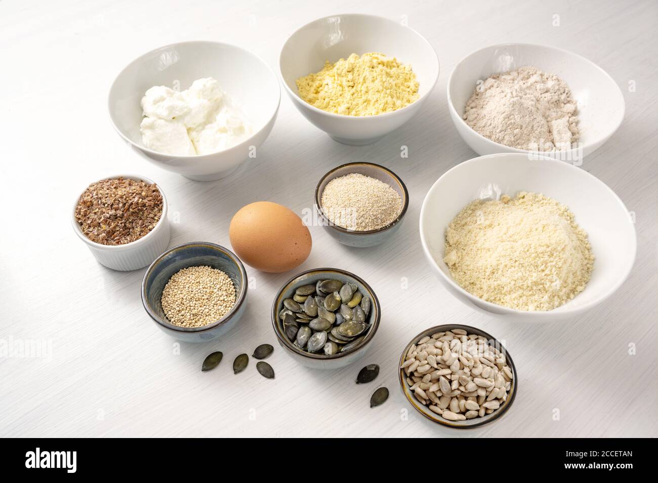 Ingrédients pour un pain aux protéines avec du fromage blanc, du son d'avoine, de la farine de lupin, de l'amande et de diverses graines dans des bols sur une table blanche, cuisson saine pour le carb bas ou Banque D'Images