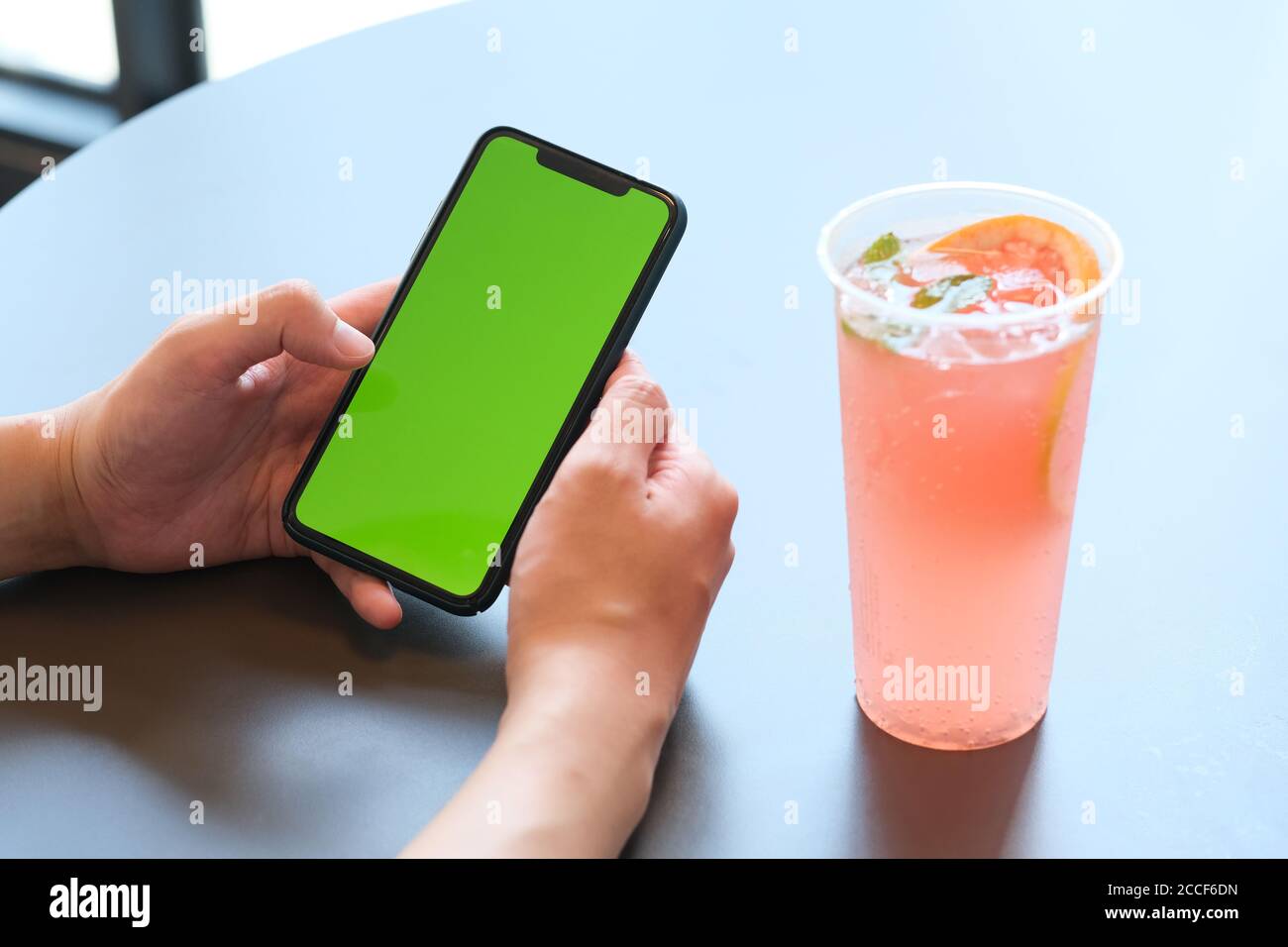 gros plan de la main tenant le smartphone à écran vert avec un une tasse de boisson froide sur une table blanche au café Banque D'Images