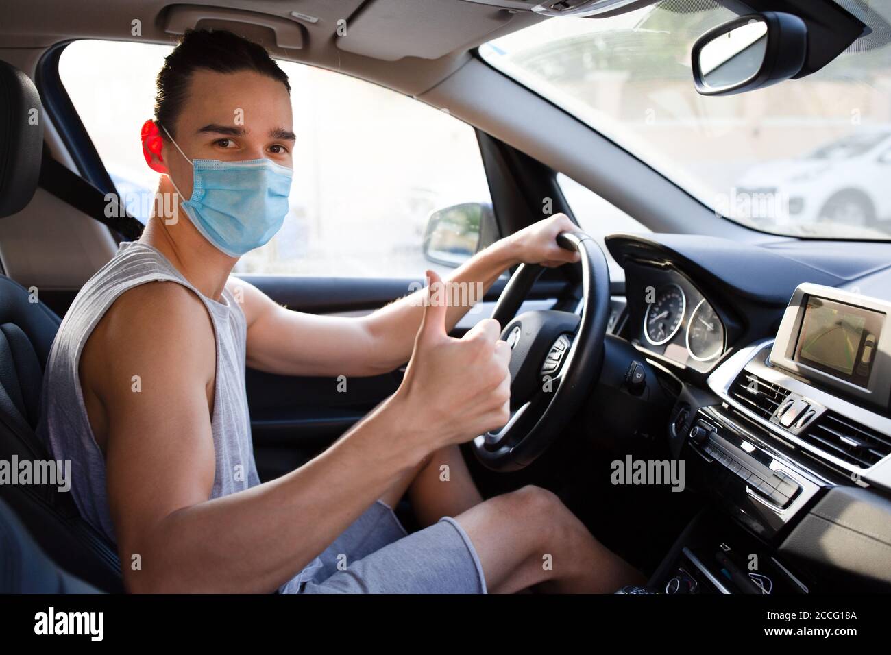Bravo ! Jeune conducteur mâle donnant un geste de main d'approbation à la caméra portant un masque médical dans la voiture. Concept de sécurité routière et de précaution. Banque D'Images