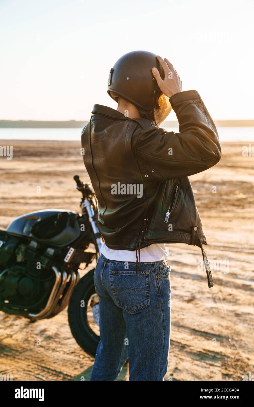 Vue arrière d'un jeune homme portant une veste en cuir et casque debout près d'une moto sur la plage ensoleillée Banque D'Images