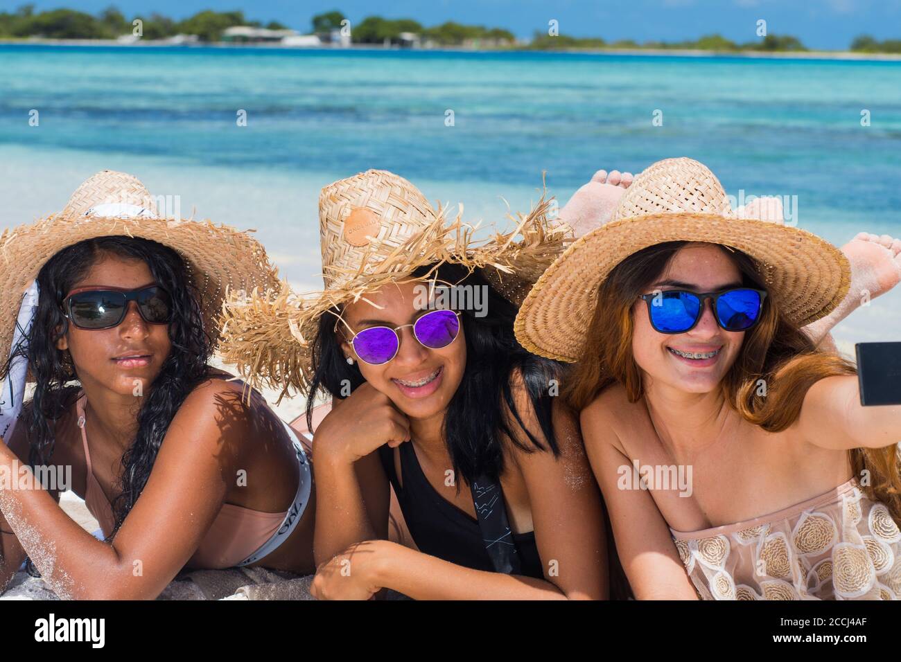 Trois adolescentes hispaniques heureux amies allongé sur une plage de sable Prendre un selfie avec un appareil photo portant un chapeau et des lunettes de soleil à Los Roques Venezuela Banque D'Images