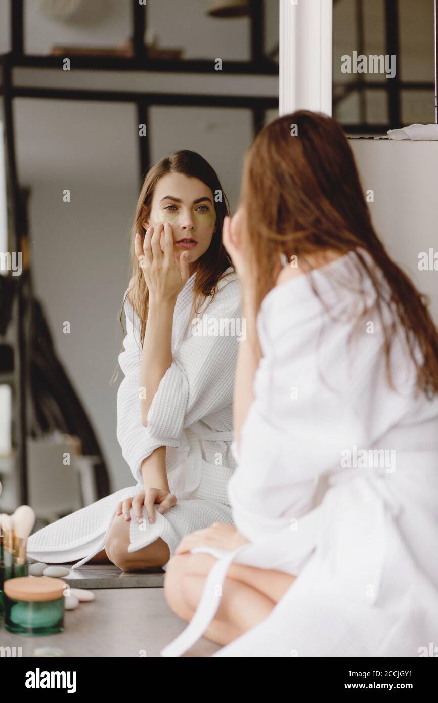 Belle jeune femme en peignoir blanc appliquant des cellules oculaires sur son visage regardant dans le miroir. Banque D'Images