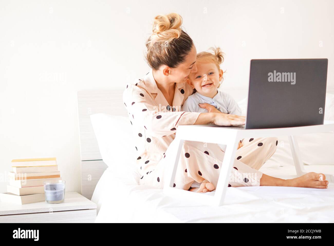 Mère et fille couchée dans le lit avec pyjama sur et Souriant les uns les autres - jeune maman travaillant de la maison dans la chambre avec son ordinateur portable sur la table du petit déjeuner Banque D'Images