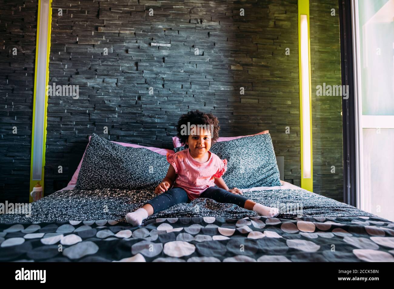 Jeune fille souriante avec les jambes écartées assis sur le lit contre mur à la maison Banque D'Images