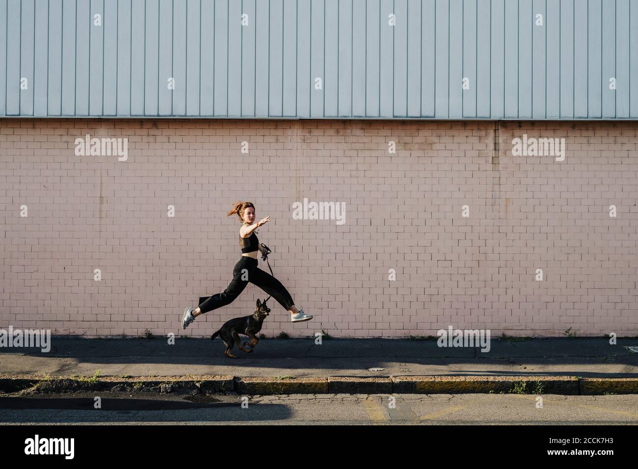 Femme qui court avec un chien sur le trottoir contre le mur Banque D'Images