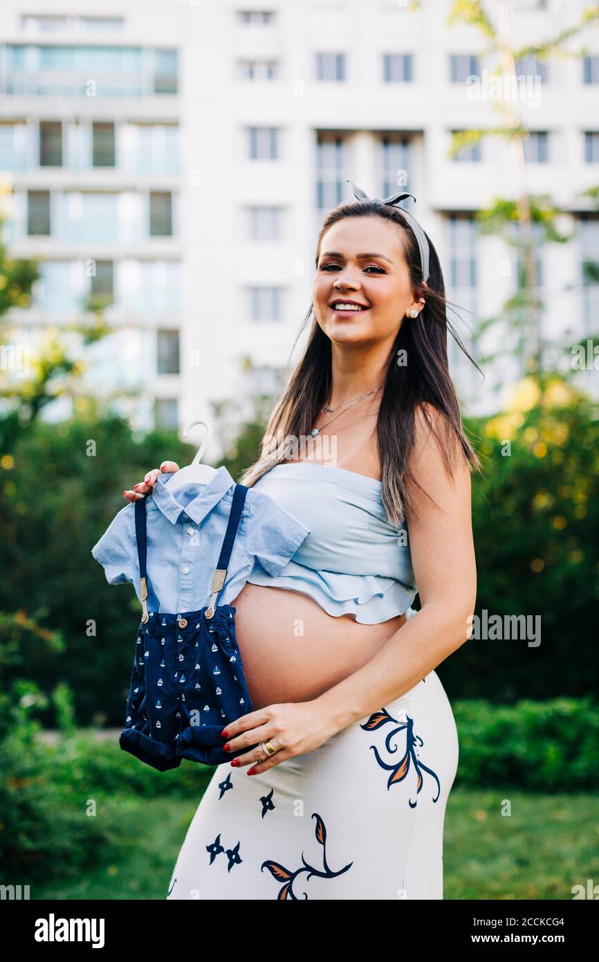 Femme enceinte souriante tenant des vêtements de bébé pendant qu'elle se trouve au parc Banque D'Images