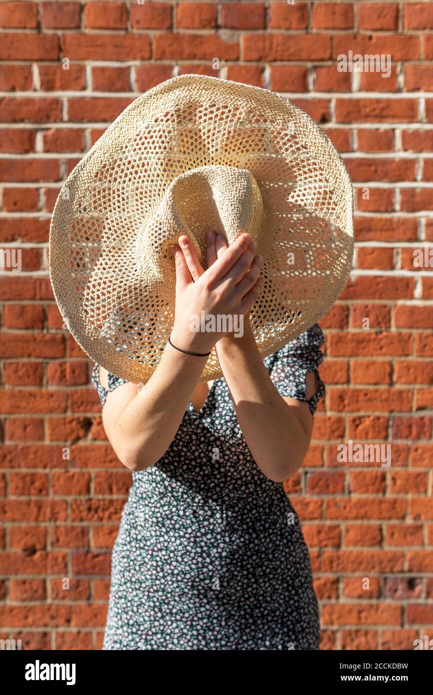 Jeune femme se cachant derrière un chapeau de paille tout en se tenant contre la brique mur pendant la journée ensoleillée Banque D'Images