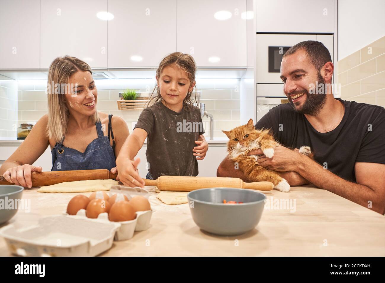 Mère et fille pétrissent de la pâte avec une goupille de pétrissage alors qu'elle était père tenir le chat sur la table de la cuisine Banque D'Images