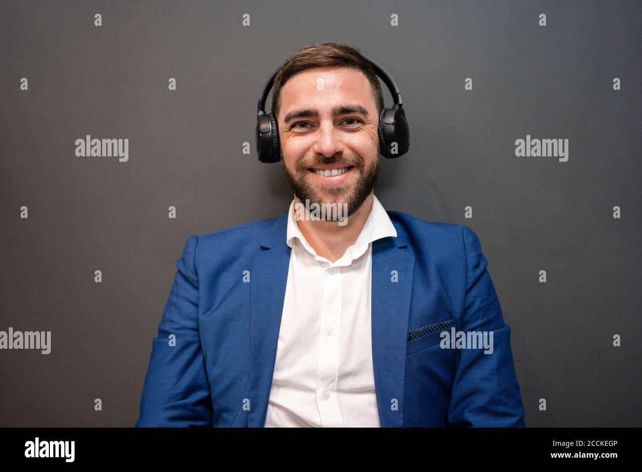 Beau homme d'affaires barbu appréciant la musique à travers des écouteurs contre le mur gris pendant la pause au bureau Banque D'Images