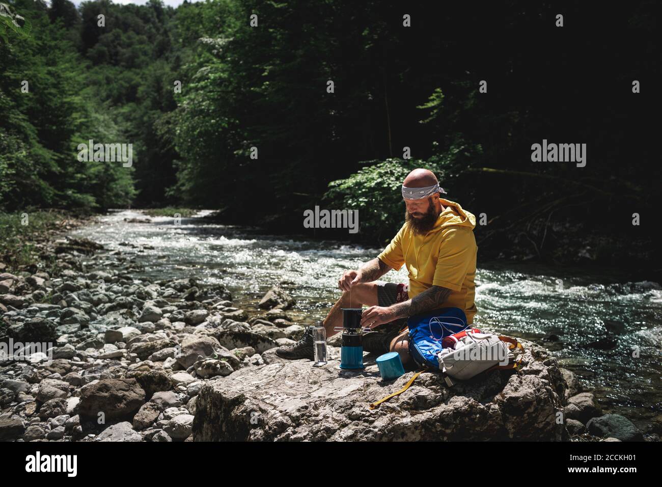 Randonneur avec une barbe complète et un sweat à capuche jaune pendant la pause, cuisant du thé au bord de la rivière Banque D'Images