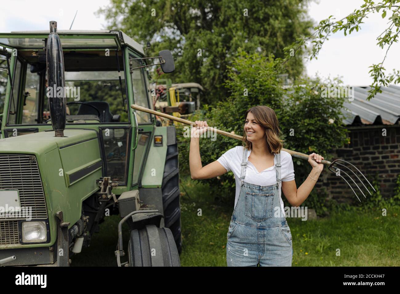 Jeune femme souriante avec fourche de foin debout à un tracteur dans la campagne Banque D'Images