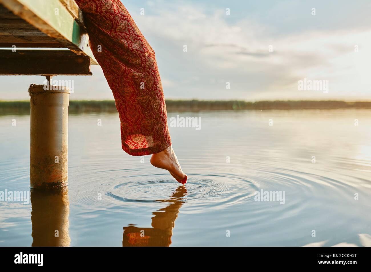 Femme assise sur la jetée d'un lac au coucher du soleil se touchant l'eau avec son pied Banque D'Images