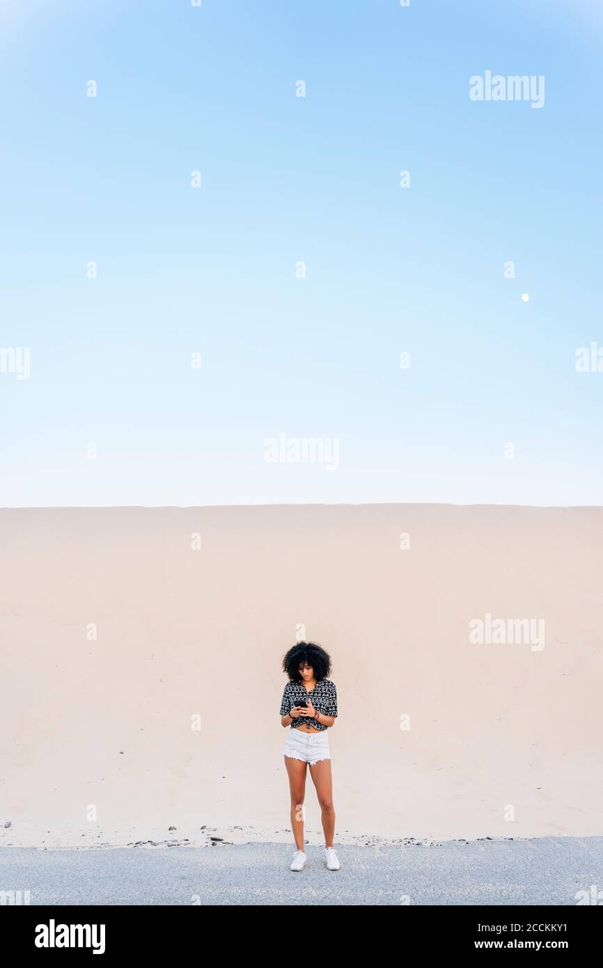 Jeune femme utilisant un téléphone portable tout en se tenant debout sur la route contre dune de sable Banque D'Images