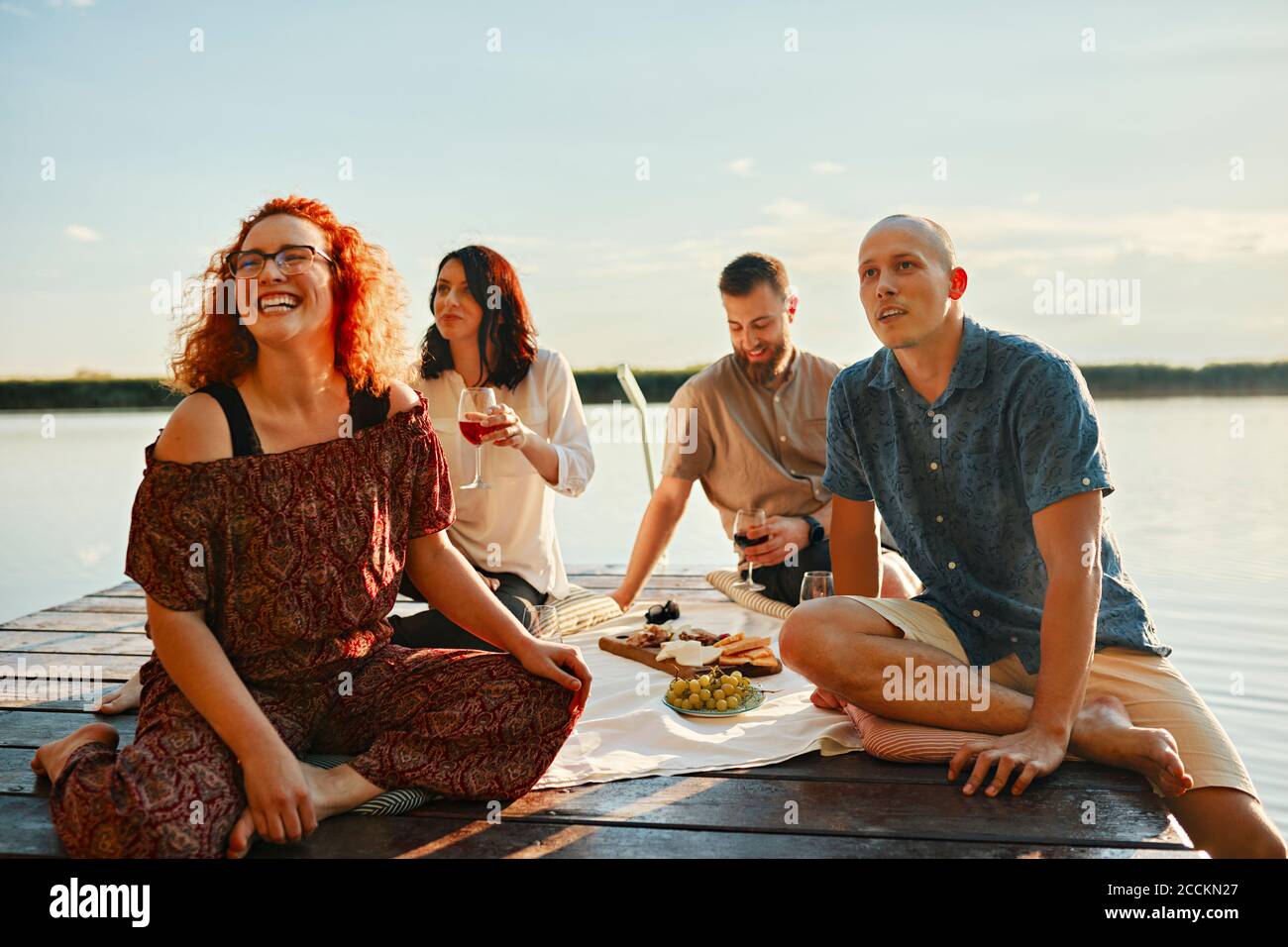 Des amis heureux ayant pique-nique sur la jetée à un lac à coucher de soleil Banque D'Images