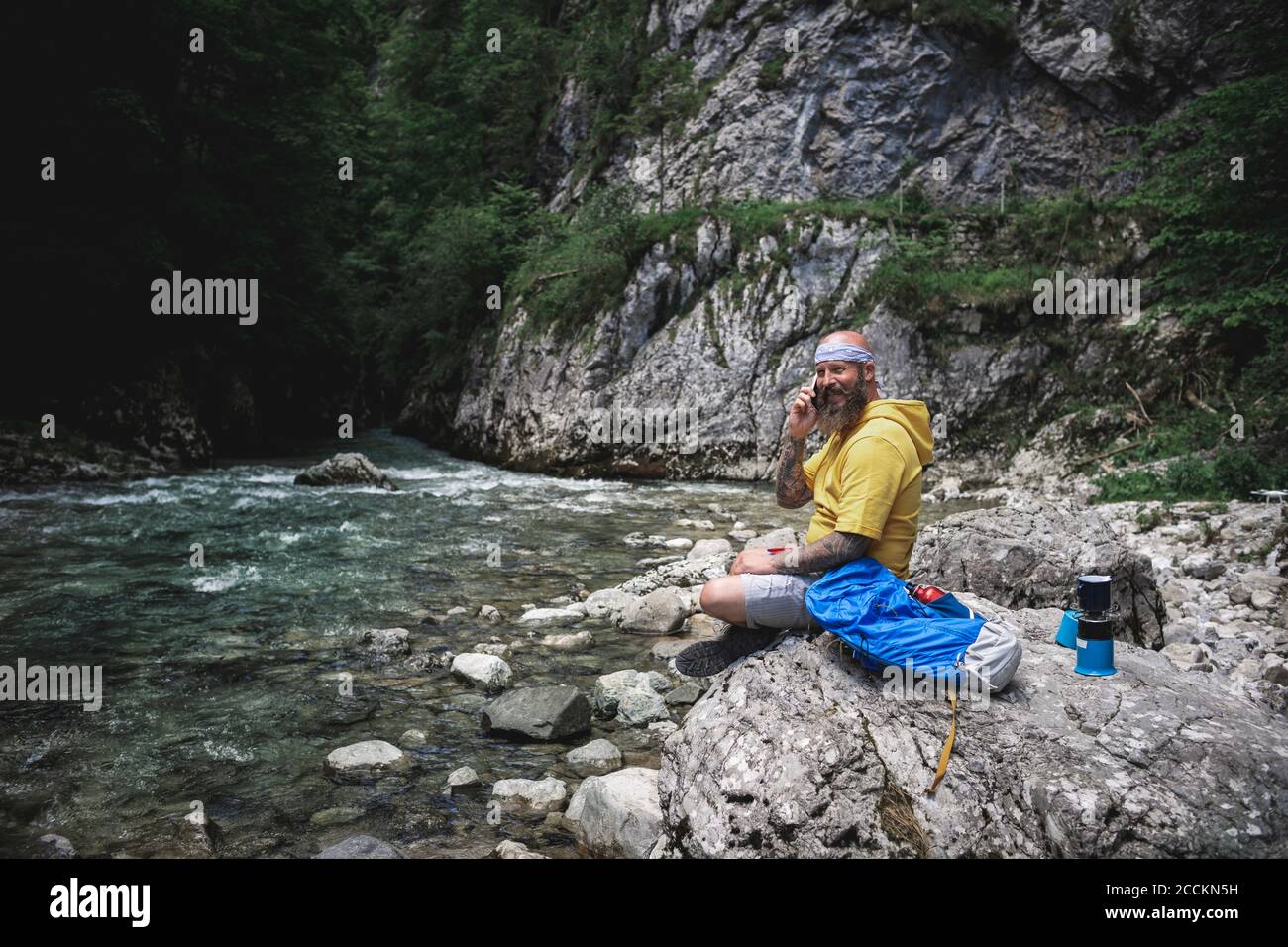 Randonneur avec une barbe complète et un sweat à capuche jaune posé sur la pierre à côté de la rivière et à l'aide d'un smartphone Banque D'Images
