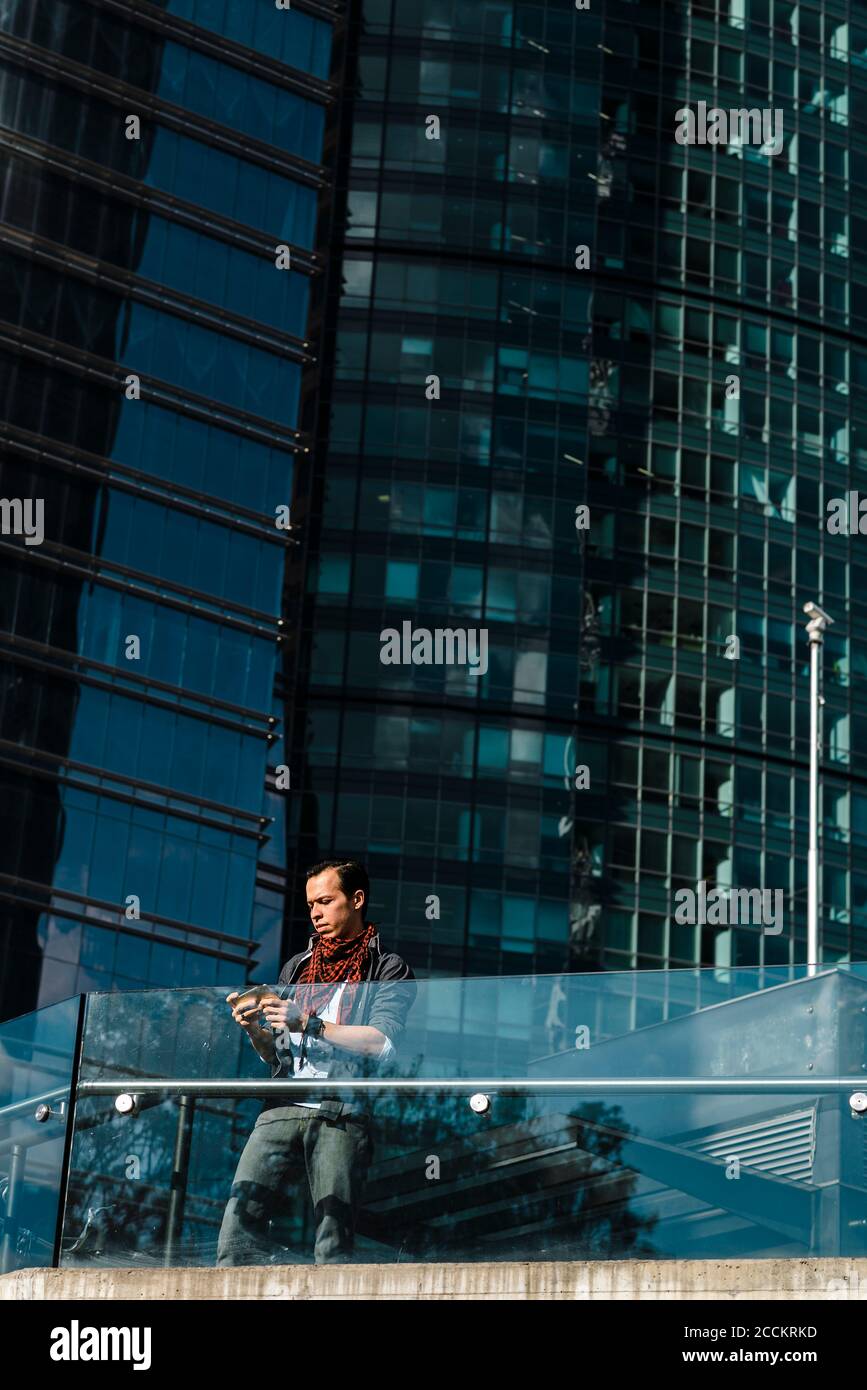 Homme utilisant un téléphone portable tout en se tenant contre un bâtiment moderne en verre en ville Banque D'Images