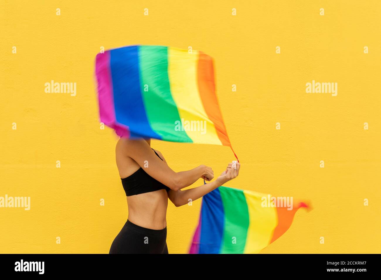 Femme agitant des drapeaux arc-en-ciel tout en se tenant contre le mur jaune à l'intérieur ville Banque D'Images