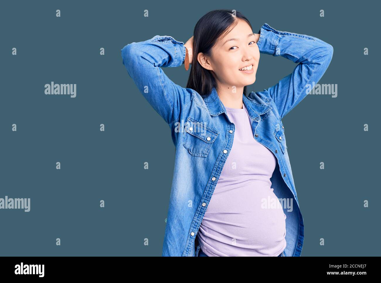 Jeune belle femme chinoise enceinte s'attendant à un bébé relaxant et étirant, bras et mains derrière la tête et le cou souriant heureux Banque D'Images