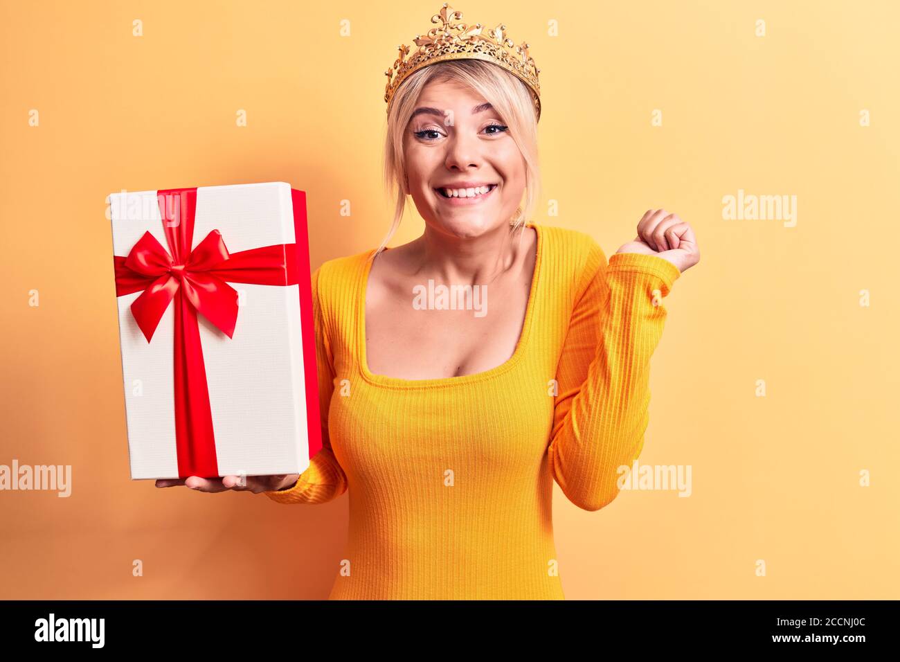 Jeune belle femme blonde portant la couronne de princesse dorée tenant le cadeau d'anniversaire criant fier, célébrant la victoire et le succès très excité avec ra Banque D'Images Jeune belle femme blonde portant la couronne de princesse dorée tenant le cadeau d'anniversaire criant fier, célébrant la victoire et le succès très excité avec ra Banque D'Images