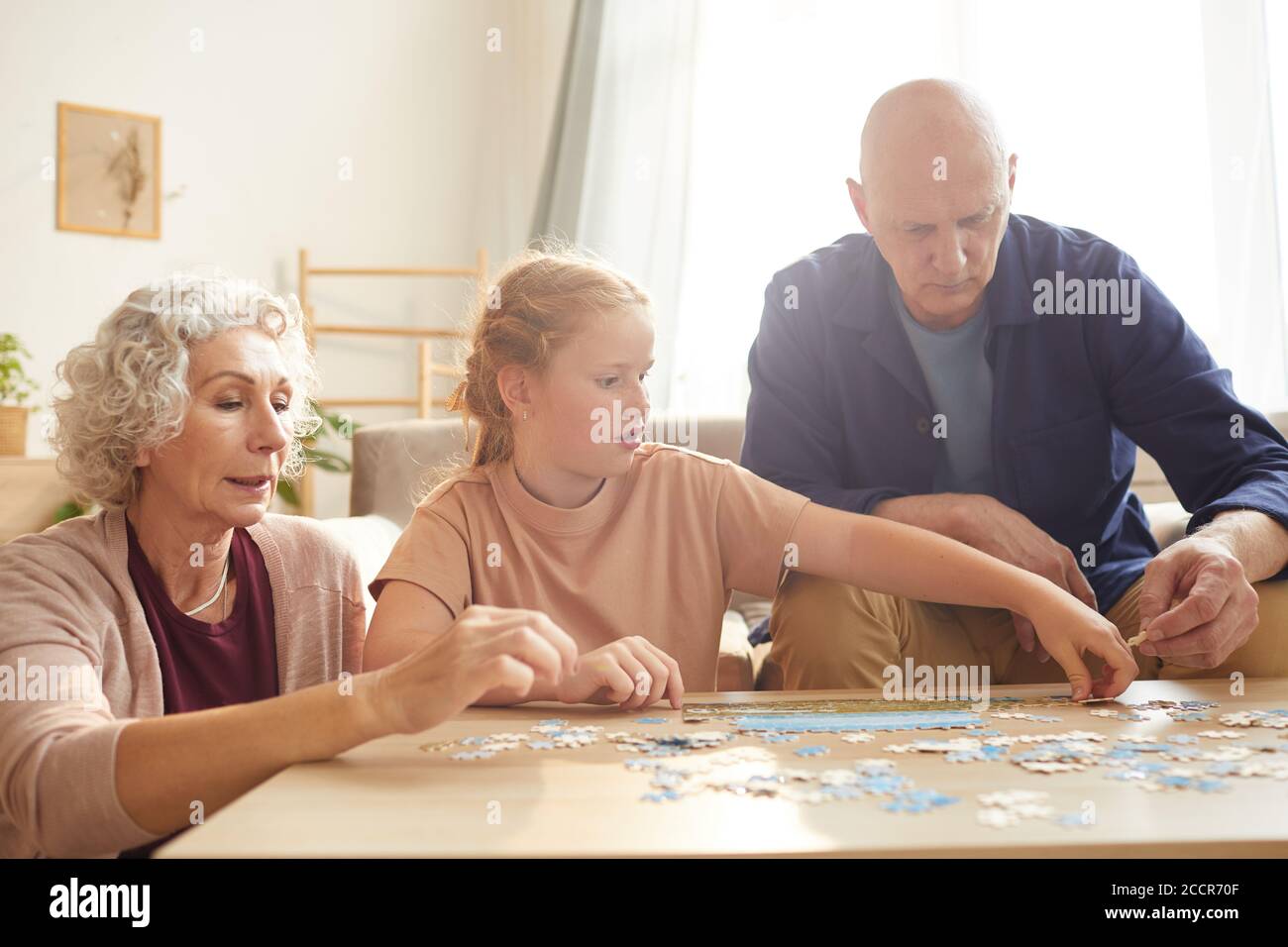Portrait de cure fille à cheveux rouges jouant à des jeux de société avec grand-parents tout en appréciant le temps ensemble dans une maison confortable éclairée par lumière du soleil Banque D'Images