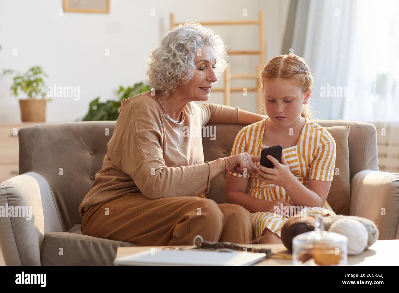Portrait aux tons chaleureux d'une femme âgée élégante montrant des photos de smartphone à la petite-fille mignonne tout en étant assis sur un canapé dans une maison confortable éclairé par la lumière du soleil Banque D'Images