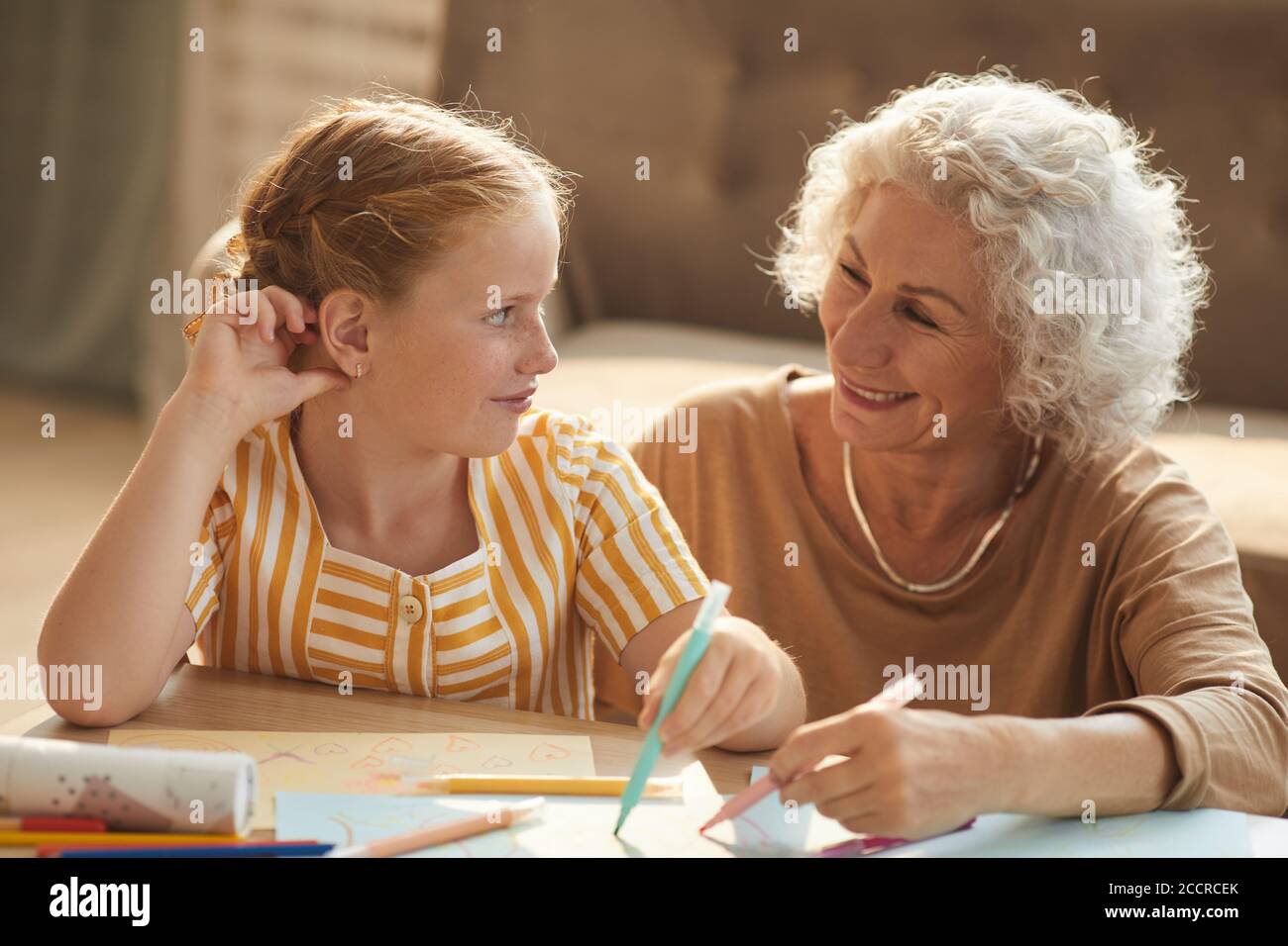 Portrait aux tons chauds d'une femme âgée souriante qui regarde une jolie fille aux cheveux rouges et qui dessine ensemble tout en étant assise sur le sol près d'une table basse dans un salon confortable. Banque D'Images