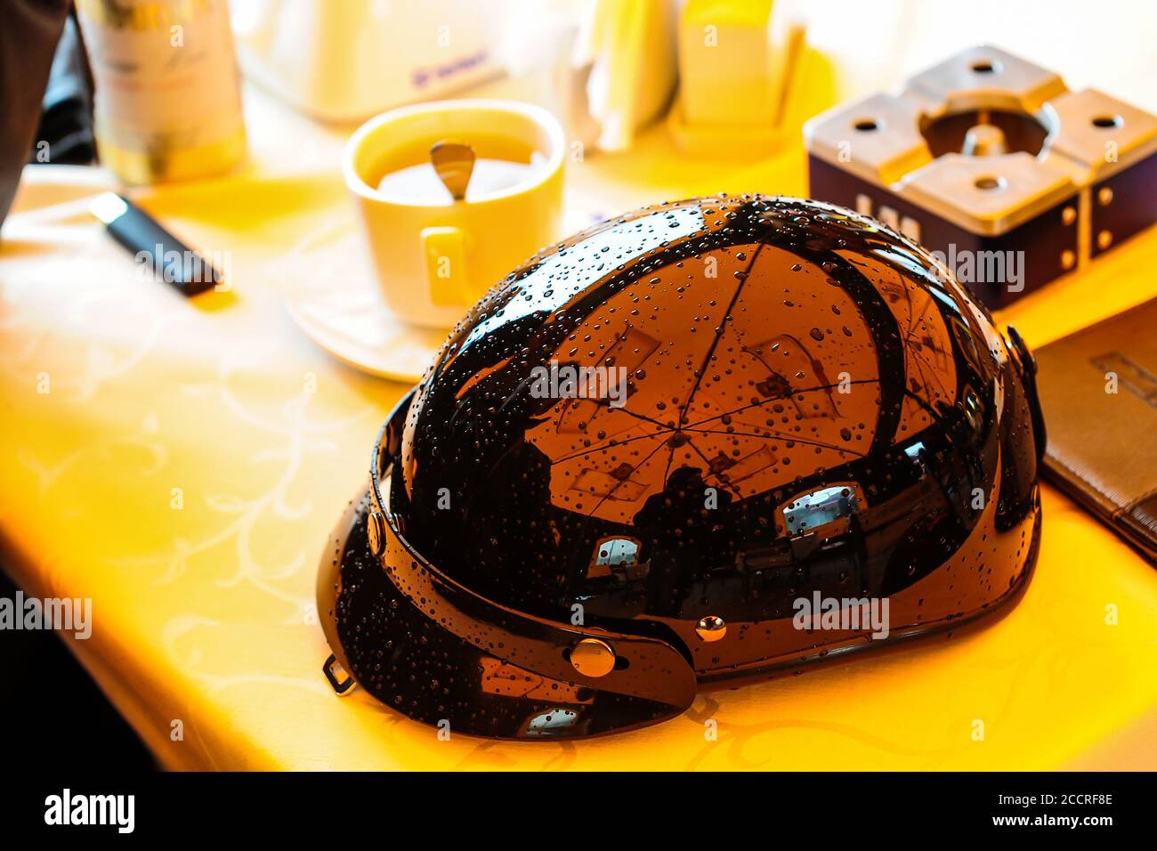 casque de moto noir brillant de style vintage avec gouttes de pluie sur une table dans un café à côté d'une tasse de café Banque D'Images