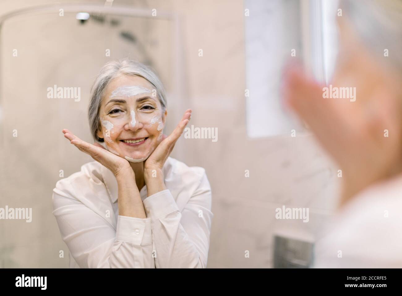 Jolie femme âgée aux cheveux gris, portant une chemise blanche, regardant  son visage dans le miroir de salle de bains, et appliquant de la crème anti- rides ou un masque cosmétique Photo Stock -
