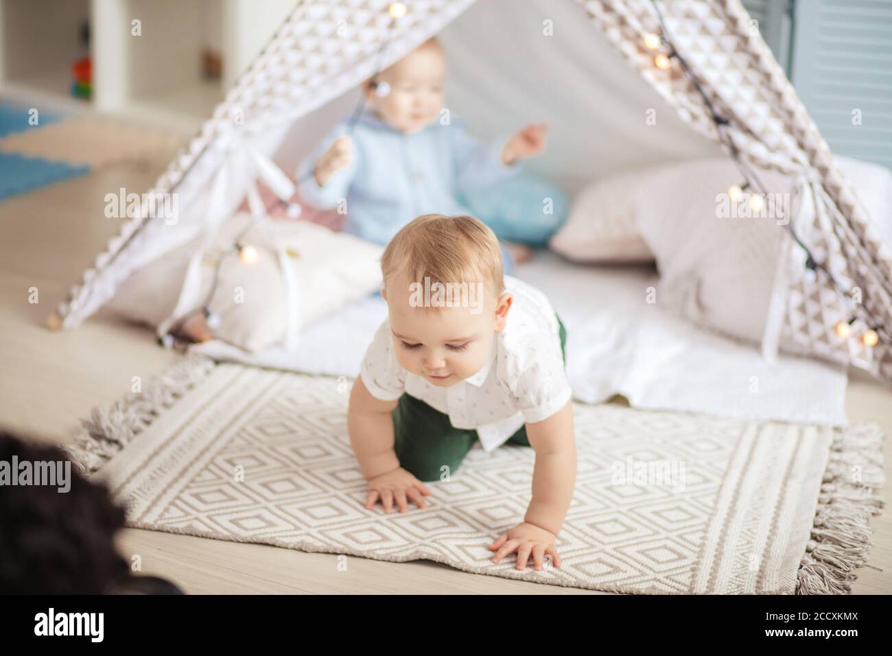 Deux jeunes enfants du même âge s'assoient sur un tapis dans une tente à tipi, s'amusent et jouent avec des guirlandes de Noël dans une chambre confortable. Banque D'Images