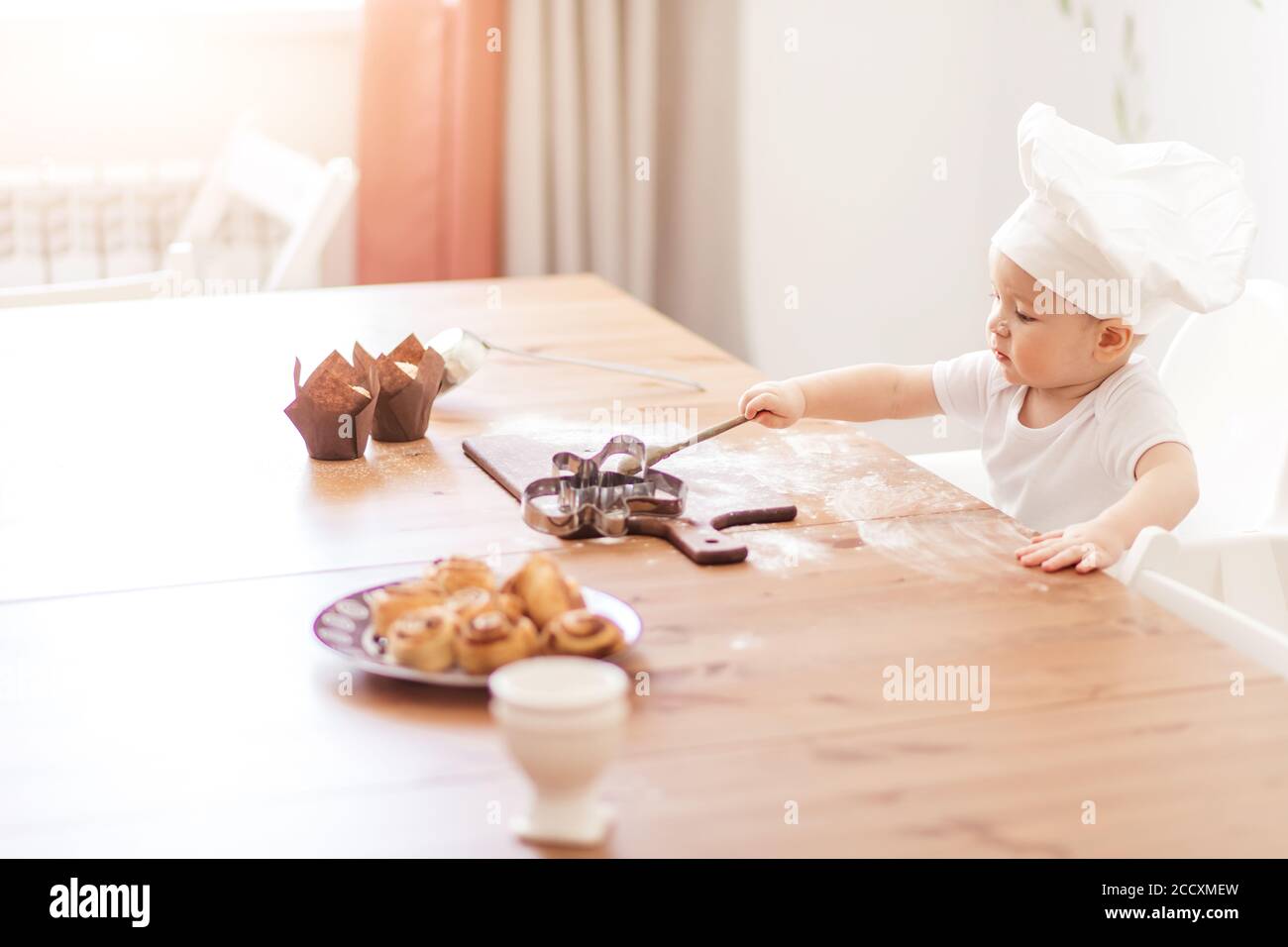 Adorable petit garçon de 1-2 ans, portant un chapeau de cuisinier jouant avec des moules à muffins, assis à une table recouverte de farine et de biscuits servis. Banque D'Images