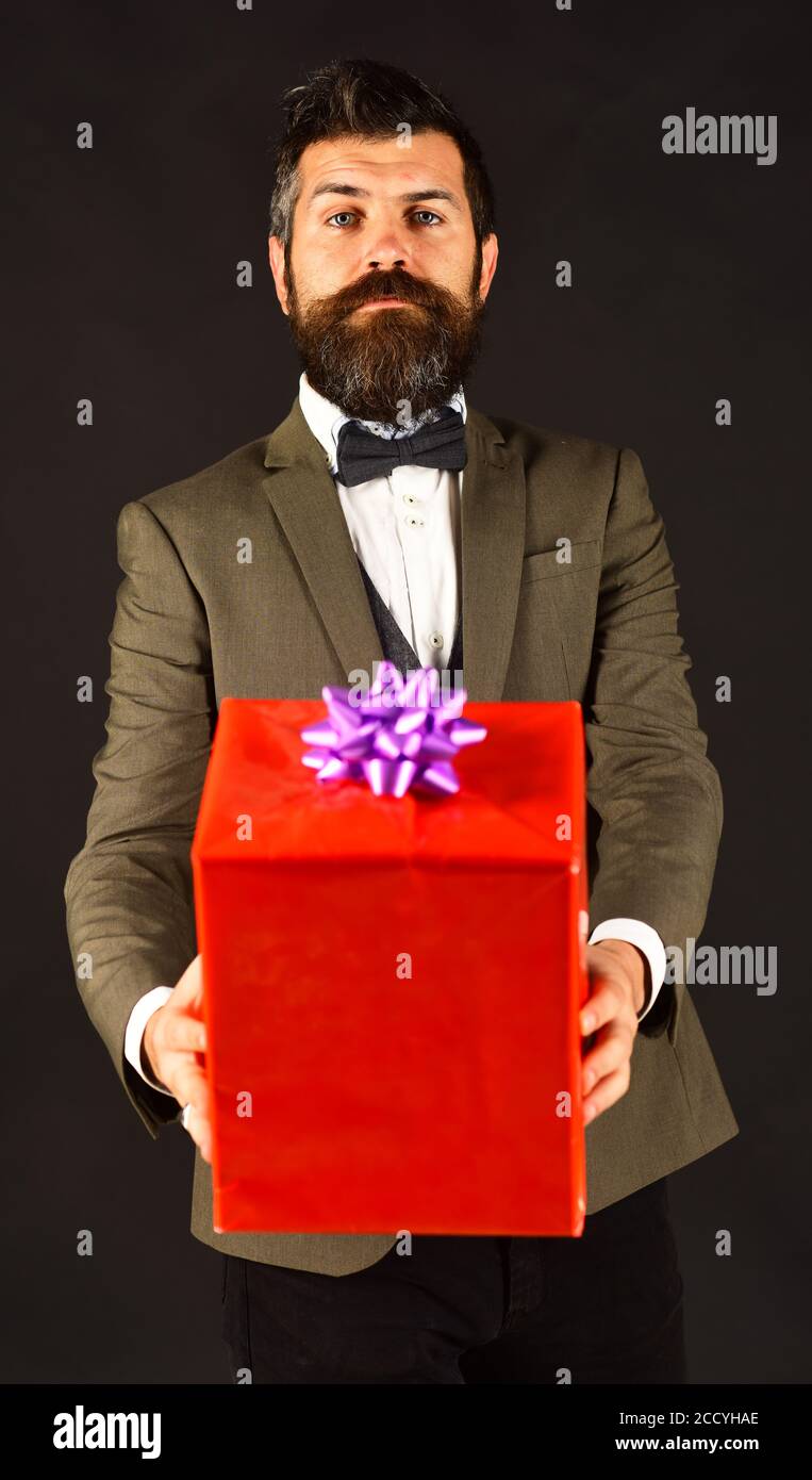 L'homme avec la barbe tient présent sur fond marron. Macho en costume rétro présente cadeau emballé. Concept cadeau anniversaire. Homme d'affaires avec visage ouvert et boîte cadeau rouge. Banque D'Images