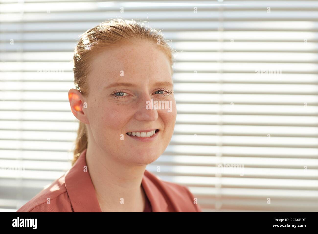 Portrait en gros plan franc d'une jeune femme aux cheveux rouges souriant à l'appareil photo tout en posant contre les blinds dans un bureau éclairé par le soleil, espace de copie Banque D'Images