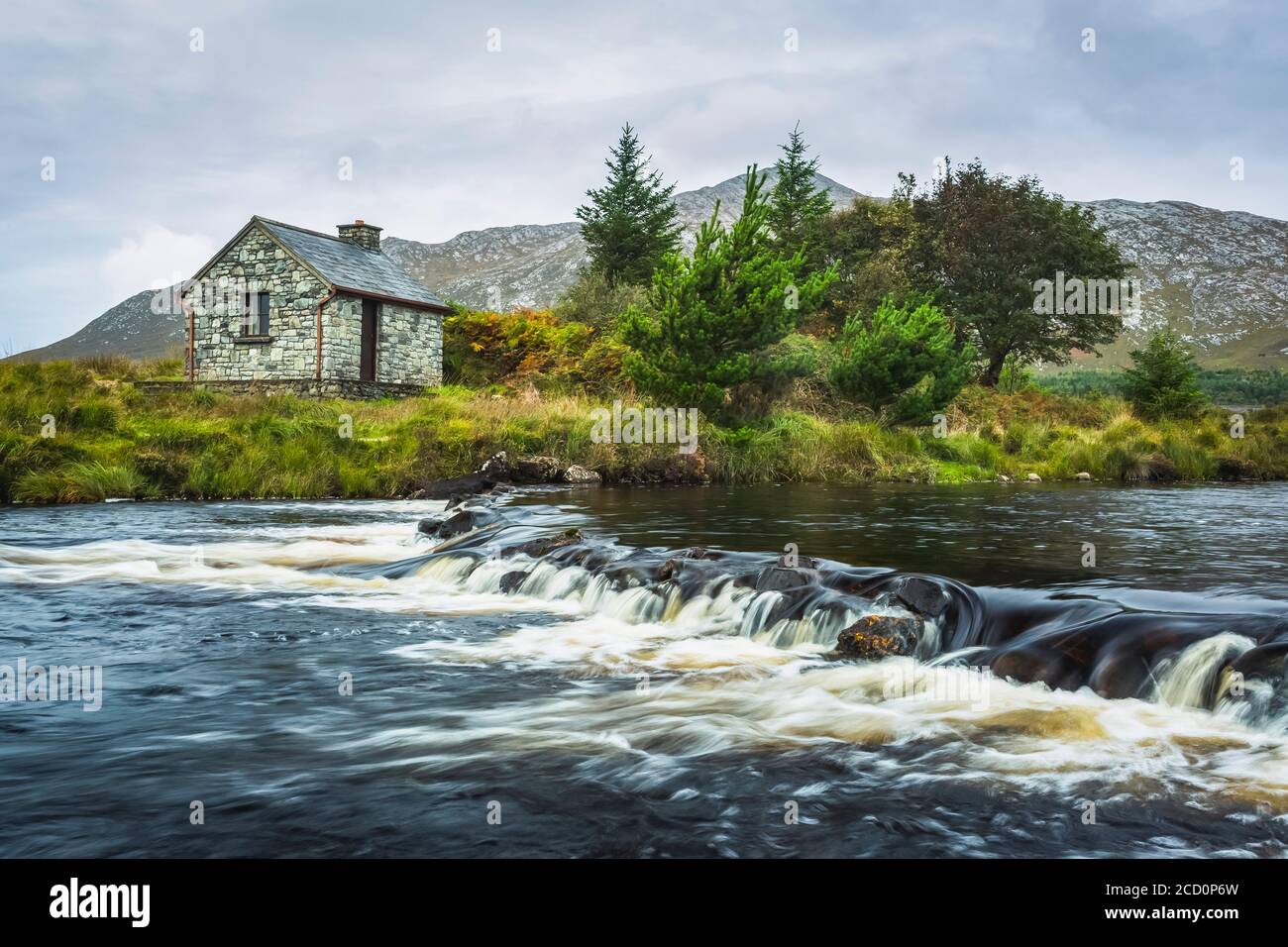 Petite cabane de pêcheur en pierre sur les rives d'une petite rivière avec des montagnes en arrière-plan par une journée nuageux; Connemara, comté de Galway, Irlande Banque D'Images