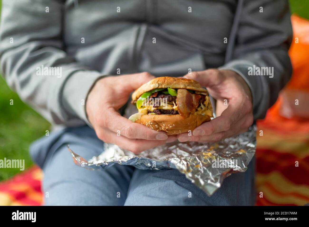 Un homme est assis sur une couverture de pique-nique avec un morceau de papier d'aluminium sur ses genoux, tenant un hamburger entre ses mains; North Vancouver, Colombie-Britannique, Canada Banque D'Images