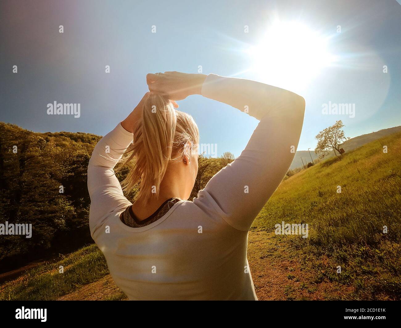 Vue arrière photo de la jeune fille blonde portant des vêtements de sport nouant une queue de cheval dans la nature. Lumière du matin. Temps ensoleillé. Mode de vie sain et sport conc Banque D'Images