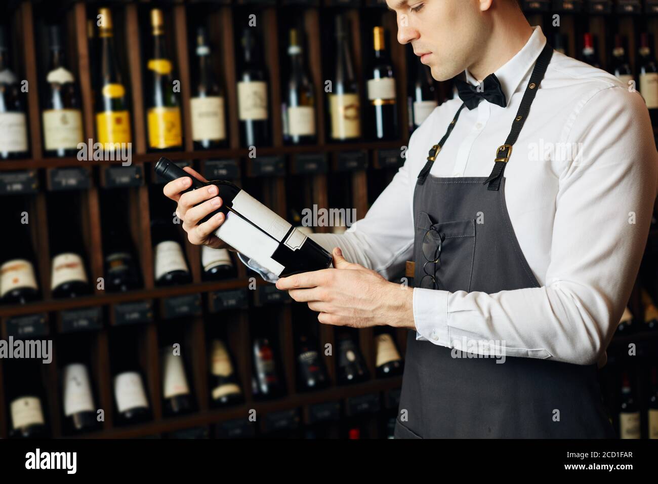 Jeune caviste caucasien vêtu d'une chemise blanche et d'un noeud papillon dans une grande boutique de vins présentant une bouteille de vin rouge au client Banque D'Images