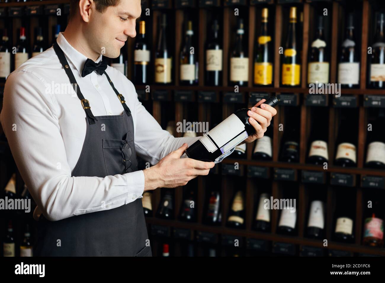 Jeune caviste caucasien vêtu d'une chemise blanche et d'un noeud papillon dans une grande boutique de vins présentant une bouteille de vin rouge au client Banque D'Images