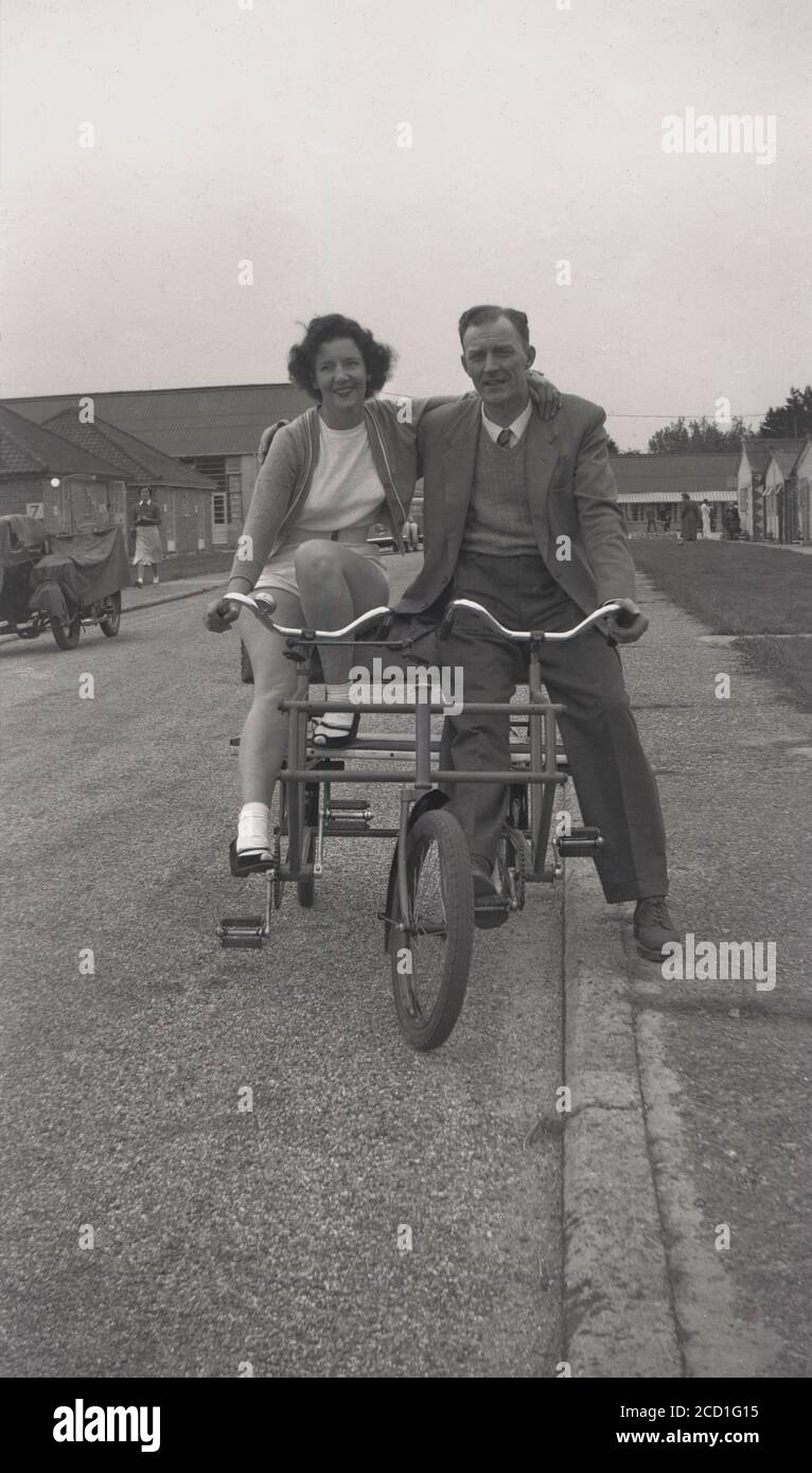 Années 1950, historique, un homme et une dame assis ensemble sur un tricycle social de deux places sur un chemin à l'extérieur des chalets dans un camp de vacances à Hayling Island, Hampshire, Angleterre, Royaume-Uni. Ces bicyclettes robustes à trois roues encadrées d'acier avec leurs sièges jumeaux et leurs poignées ont permis à deux amis ou membres de la famille de s'asseoir côte à côte en faisant du vélo et ont été une forme de transport populaire et amusant pour se déplacer dans la grande étendue d'un camp de vacances dans cette période d'après-guerre. Banque D'Images