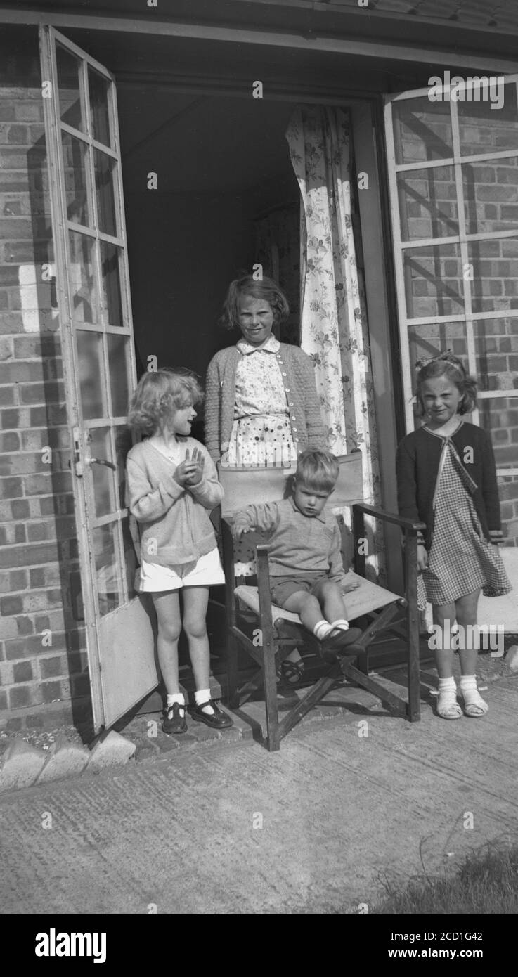 Dans les années 1950, historique, trois jeunes filles et un garçon de jeu à l'extérieur des portes en métal français de leur chalet dans un camp de vacances à Hayling Island, Hampshire, Angleterre, Royaume-Uni, avec le petit garçon assis dans la chaise! Banque D'Images