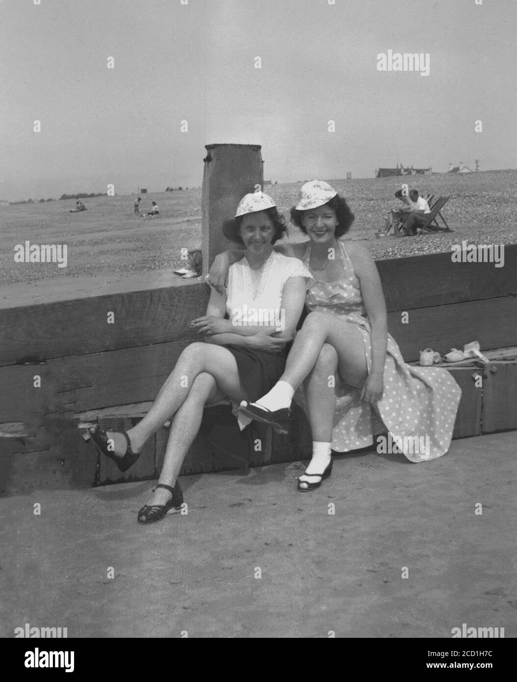 années 1950, historique, « une jambe de force » ! Deux dames assises sur un brise-roche en bois à la plage avec leurs jupes tirées vers le haut pour obtenir un peu de soleil sur leurs jambes, Hayling Island, Angleterre, Royaume-Uni. L'une des femmes porte des chaussettes et des chaussures blanches de longueur cheville, un look populaire parmi les femmes de cette époque. Banque D'Images