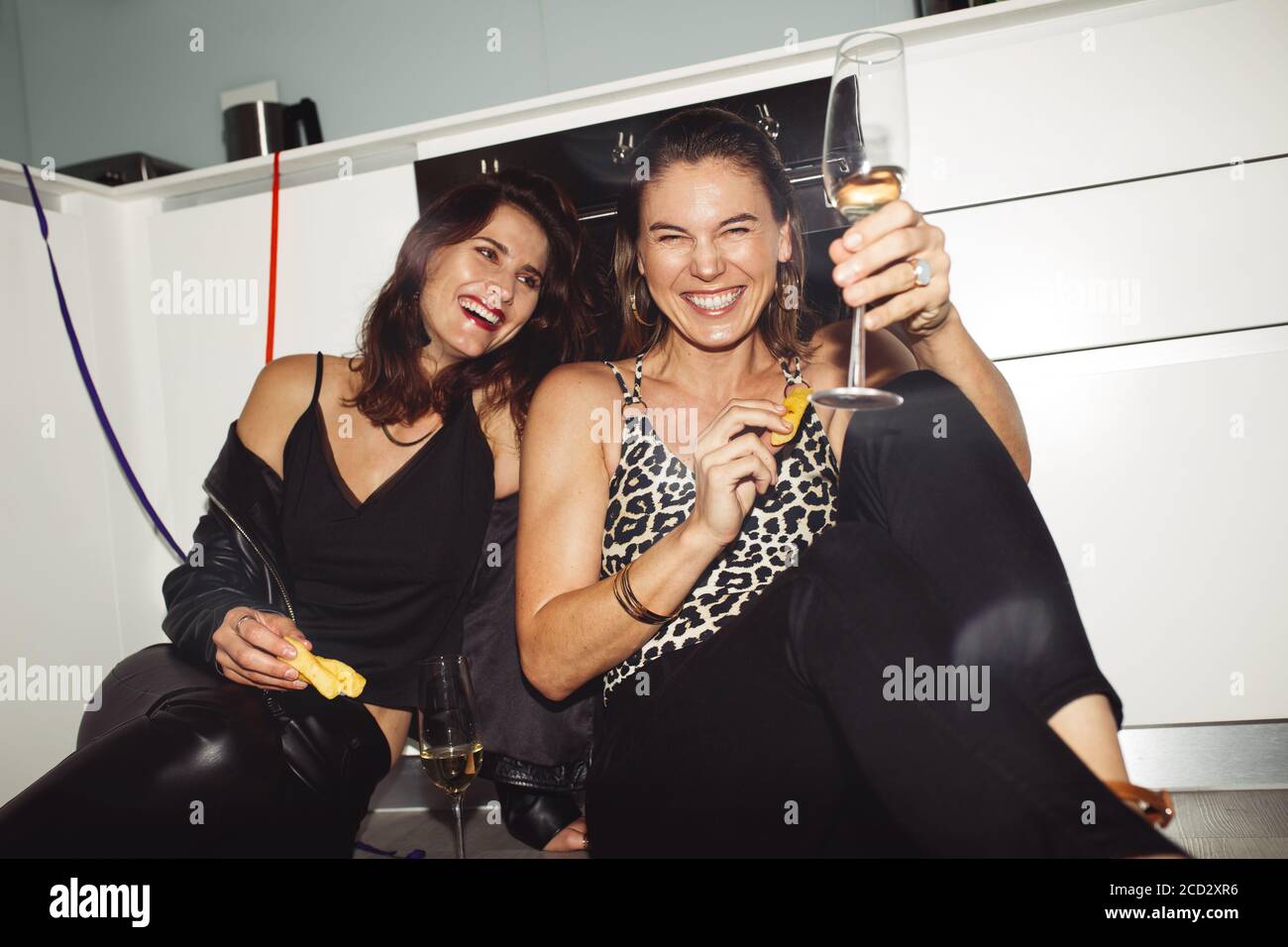 Des femmes heureuses qui s'amusent à la maison en dégustant du vin. Deux femmes gaies ayant une fête assise sur le sol à la maison. Banque D'Images