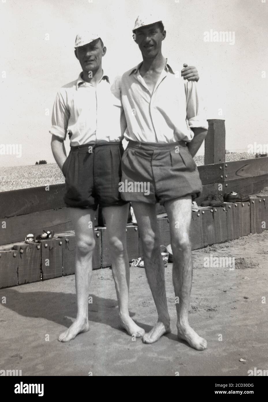 Dans les années 1950, historiques, deux hommes se tenant ensemble pour une  photo sur une plage de sable dans leurs pieds, avec des casquettes blanches  sur leur tête, des manches enroulées les