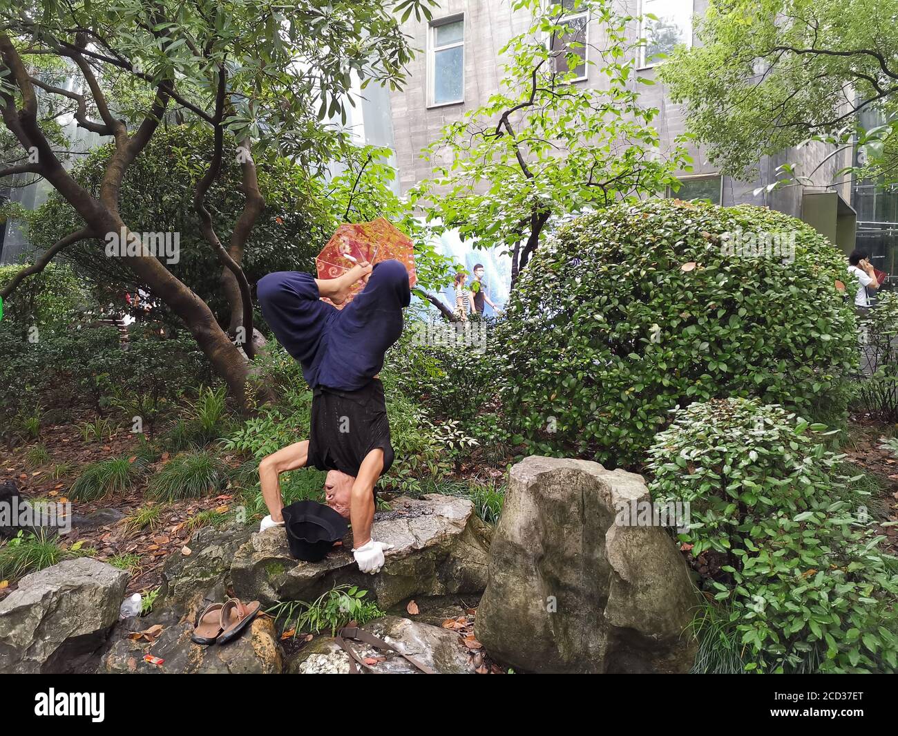 M. Jiang, un homme dans ses années soixante-dix, pratique le yoga d'une manière spéciale, debout à l'envers avec sa tête tout en tenant un parapluie avec son pied sur un ro Banque D'Images