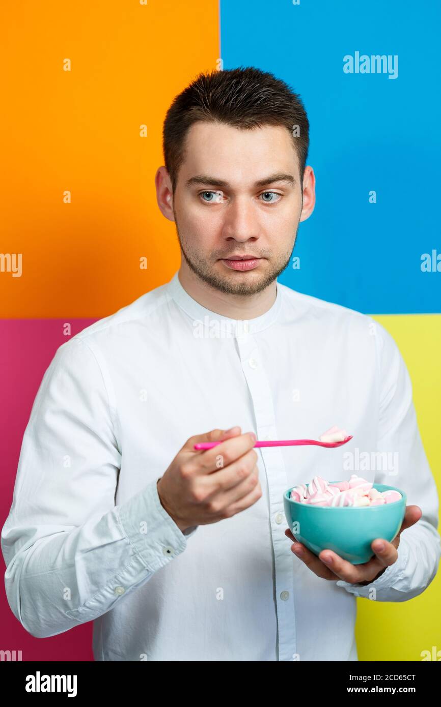 Un homme sur fond jaune, bleu, orange, rose dans une chemise blanche avec un plateau turquoise de guimauves et une cuillère rose. Le gars avec un sourire. Un im Banque D'Images